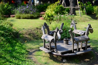 A wooden merry-go-round with four carved horse seats is situated in a lush, green garden. The play structure is surrounded by well-maintained grass and various flowering plants, creating a serene and playful atmosphere.