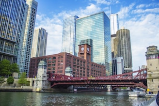 A cityscape featuring a skyline of modern glass skyscrapers and a historic red brick building with a clock tower. A red drawbridge spans a river, with reflections shimmering on the water. The sky is partly cloudy, creating a bright and inviting atmosphere.