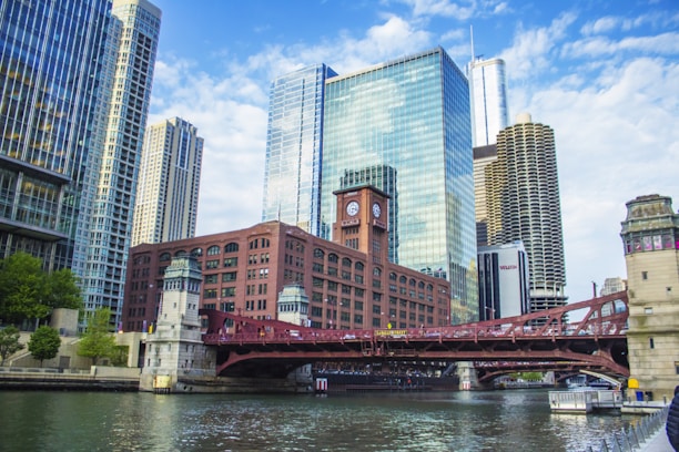 A cityscape featuring a skyline of modern glass skyscrapers and a historic red brick building with a clock tower. A red drawbridge spans a river, with reflections shimmering on the water. The sky is partly cloudy, creating a bright and inviting atmosphere.