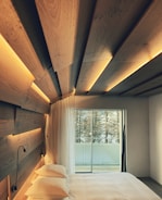 A bedroom featuring a modern design with wood paneling on the ceiling and walls, softly illuminated by integrated lights. A large glass sliding door opens up to a view of a balcony and trees outside. The room contains a neatly made bed with white bedding and dual reading lamps attached to the wooden headboard.
