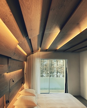 A bedroom featuring a modern design with wood paneling on the ceiling and walls, softly illuminated by integrated lights. A large glass sliding door opens up to a view of a balcony and trees outside. The room contains a neatly made bed with white bedding and dual reading lamps attached to the wooden headboard.