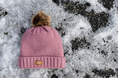 A knitted pink winter hat with a faux fur pom-pom on top is placed on the snowy ground. The hat features a branded label on the front. The background consists of a patchy layer of snow with dark, icy patches.