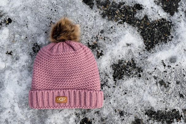 A knitted pink winter hat with a faux fur pom-pom on top is placed on the snowy ground. The hat features a branded label on the front. The background consists of a patchy layer of snow with dark, icy patches.