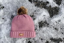 A knitted pink winter hat with a faux fur pom-pom on top is placed on the snowy ground. The hat features a branded label on the front. The background consists of a patchy layer of snow with dark, icy patches.