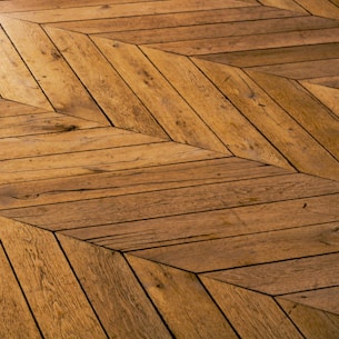 A detailed view of a wooden floor with a geometric chevron pattern. The wood planks are arranged in a herringbone style, showcasing various shades and textures of brown.