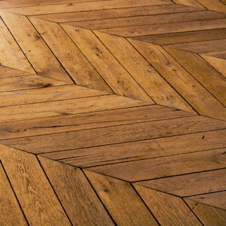 A detailed view of a wooden floor with a geometric chevron pattern. The wood planks are arranged in a herringbone style, showcasing various shades and textures of brown.