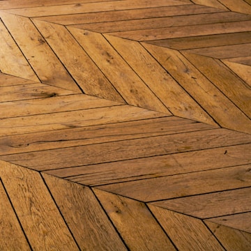 A detailed view of a wooden floor with a geometric chevron pattern. The wood planks are arranged in a herringbone style, showcasing various shades and textures of brown.