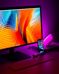 A modern desk setup featuring a large desktop monitor displaying a colorful abstract wallpaper with vibrant orange and blue waves. Next to the monitor, an iPhone with applications visible on the home screen is placed upright on a stand. The desk is illuminated by a soft purple light, creating a vibrant and high-tech atmosphere. A wireless keyboard sits in front of the monitor, and a small device, likely AirPods, is beside the phone.