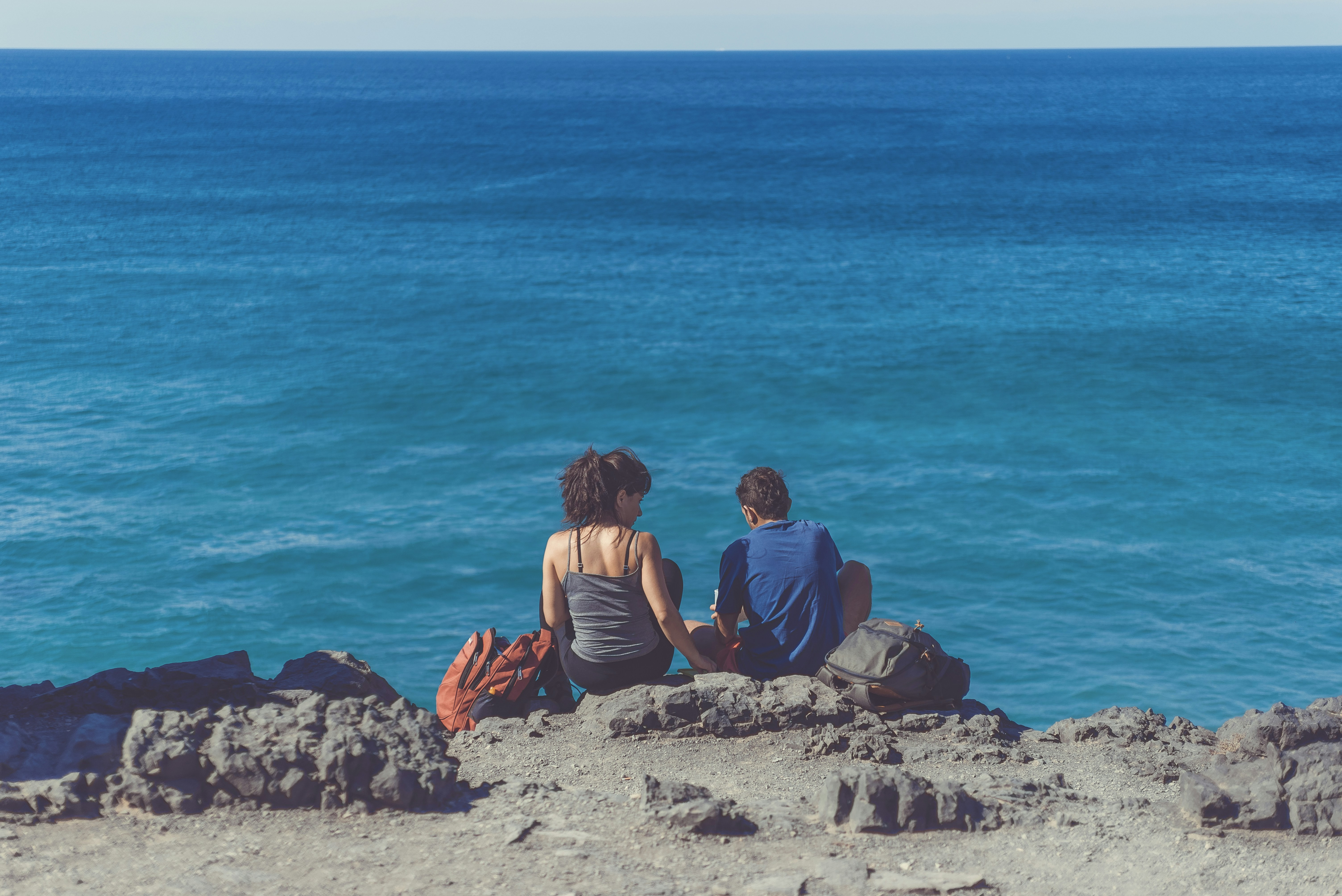man and woman sitting next to each other on cliff in front of body of water