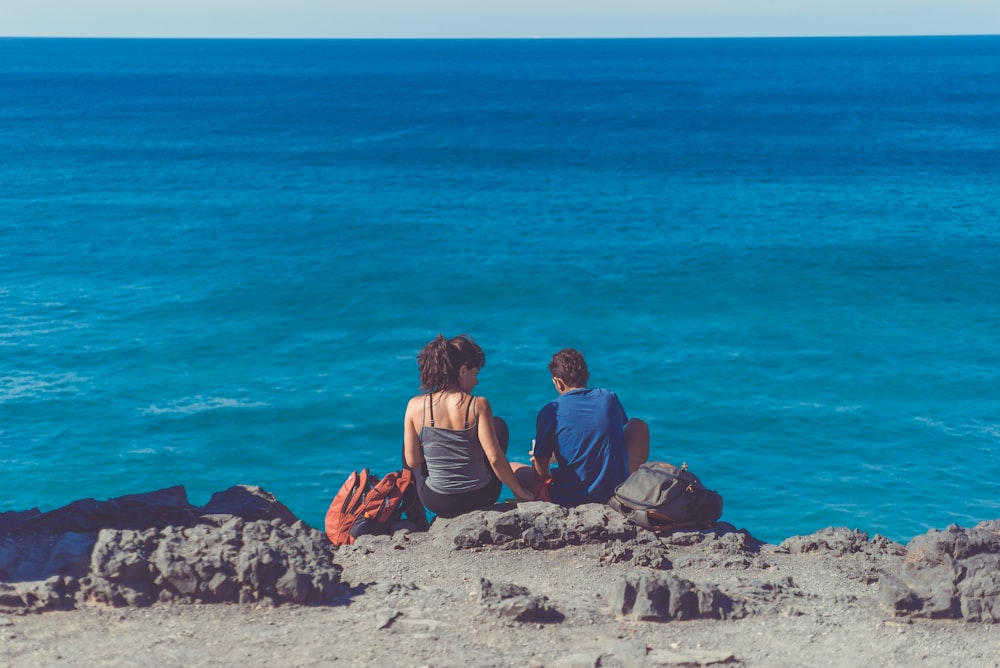 man and woman sitting next to each other on cliff in front of body of water