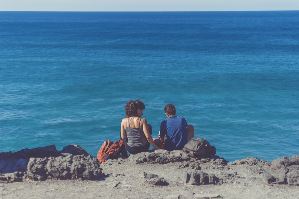 man and woman sitting next to each other on cliff in front of body of water