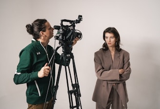 A woman wearing a brown suit stands with her arms crossed next to a man operating a professional video camera on a tripod. The setting appears to be a studio with a neutral background.
