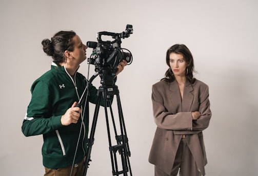 A woman wearing a brown suit stands with her arms crossed next to a man operating a professional video camera on a tripod. The setting appears to be a studio with a neutral background.