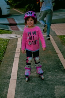 A young child wearing a pink shirt with the words 'PLAY ALL DAY', a pink helmet, knee pads, and rollerblades skates on a paved path. In the background, an adult in casual attire is walking away, and there are some green grassy areas and a white car further back.