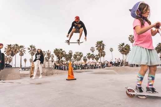 A skateboarder performs a high jump trick in a vibrant outdoor skatepark, while onlookers and other skaters enjoy the activity. A young girl in a colorful outfit rides a scooter in the foreground, adding a playful element to the scene. Palm trees and a crowd of spectators create a lively and sunny atmosphere.