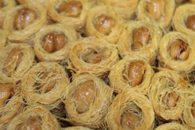 Nest-like pastries filled with nuts, intricately woven with a thread-like pastry. The texture is crispy and golden brown, with glistening nuts peeking through the center.