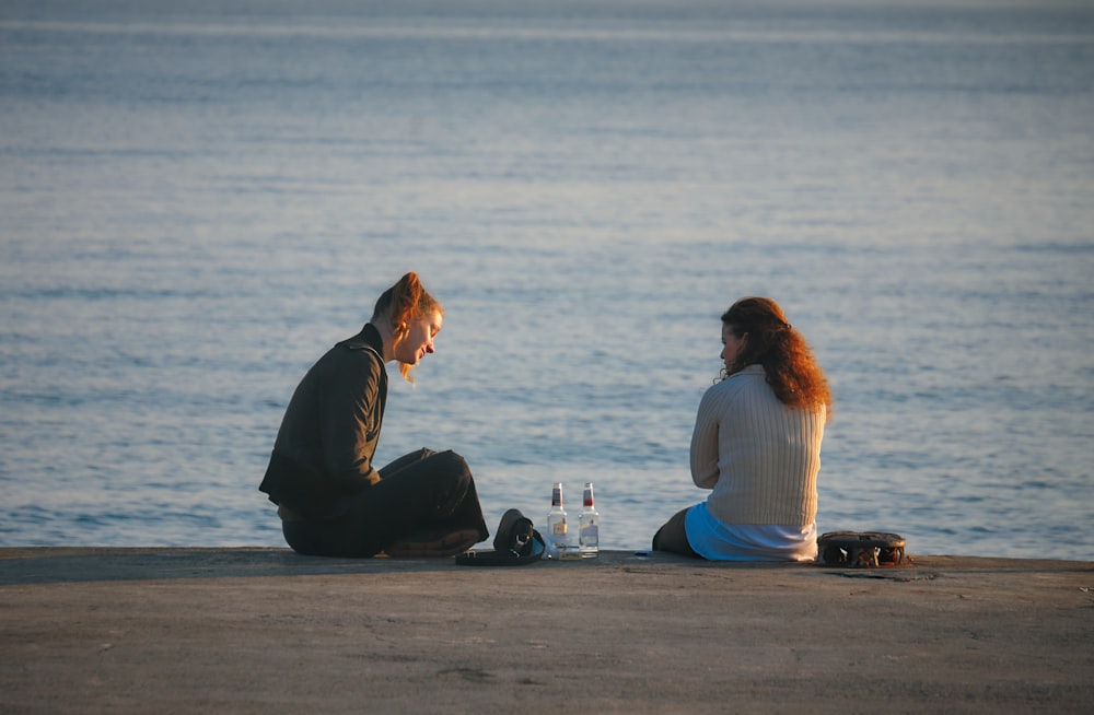 2 women sitting on concrete with bottles in between them during daytime