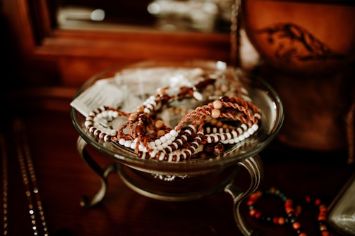 A collection of beaded bracelets rests in a decorative glass bowl on a wooden surface. The bracelets feature white and brown beads, with some incorporating reddish-brown woven elements. The background includes blurred hints of additional jewelry and a wooden object.