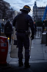 A person in riot gear stands on a city street holding a clear shield. The surroundings include other pedestrians walking past and urban architecture. The sky is cloudy, indicating an overcast day.