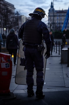 A person in riot gear stands on a city street holding a clear shield. The surroundings include other pedestrians walking past and urban architecture. The sky is cloudy, indicating an overcast day.