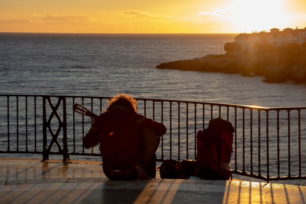 man sitting near steel railing playing guitar during golden hour