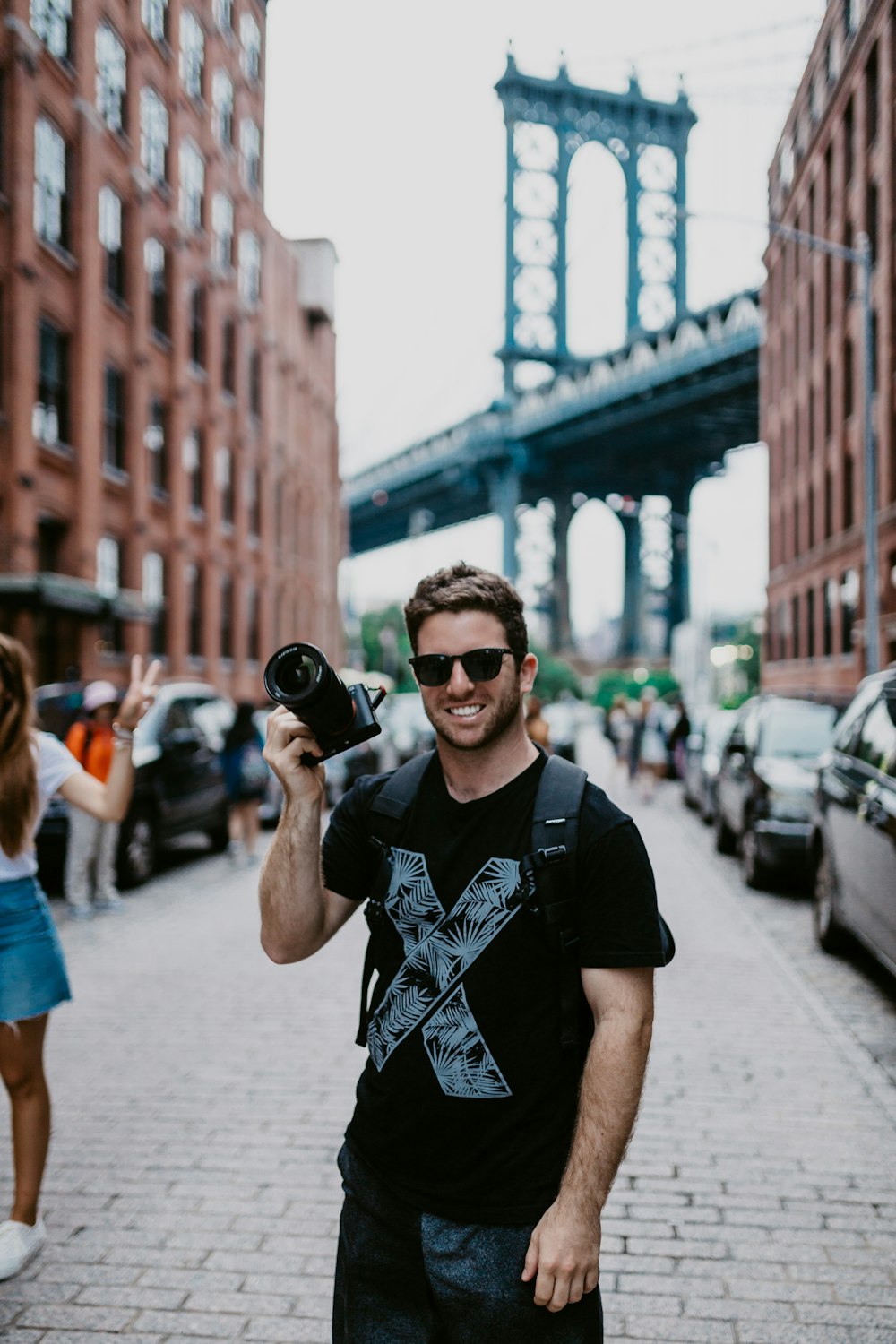 uomo sorridente che tiene lo zaino per il trasporto della fotocamera DSLR vicino alle auto parcheggiate durante il giorno