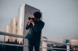 A person stands on a rooftop railing, holding a camera up to their face as they take a photograph. Behind them, a city skyline is visible with a modern glass building reflecting the soft light. The sky appears overcast, casting a moody ambiance over the scene.