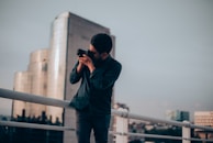 A person stands on a rooftop railing, holding a camera up to their face as they take a photograph. Behind them, a city skyline is visible with a modern glass building reflecting the soft light. The sky appears overcast, casting a moody ambiance over the scene.