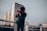 A person stands on a rooftop railing, holding a camera up to their face as they take a photograph. Behind them, a city skyline is visible with a modern glass building reflecting the soft light. The sky appears overcast, casting a moody ambiance over the scene.