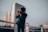 A person stands on a rooftop railing, holding a camera up to their face as they take a photograph. Behind them, a city skyline is visible with a modern glass building reflecting the soft light. The sky appears overcast, casting a moody ambiance over the scene.