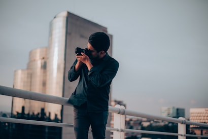 A person stands on a rooftop railing, holding a camera up to their face as they take a photograph. Behind them, a city skyline is visible with a modern glass building reflecting the soft light. The sky appears overcast, casting a moody ambiance over the scene.