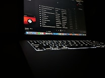 A darkly lit scene featuring a laptop with a glowing keyboard and a screen displaying a music streaming application. The interface shows a playlist with visible song titles and album artwork. Application icons are aligned at the bottom of the screen, indicating a computer interface.