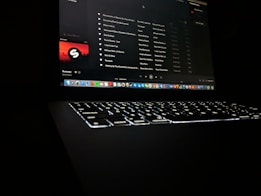 A darkly lit scene featuring a laptop with a glowing keyboard and a screen displaying a music streaming application. The interface shows a playlist with visible song titles and album artwork. Application icons are aligned at the bottom of the screen, indicating a computer interface.