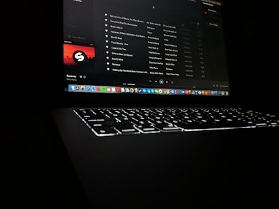 A darkly lit scene featuring a laptop with a glowing keyboard and a screen displaying a music streaming application. The interface shows a playlist with visible song titles and album artwork. Application icons are aligned at the bottom of the screen, indicating a computer interface.