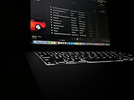 A darkly lit scene featuring a laptop with a glowing keyboard and a screen displaying a music streaming application. The interface shows a playlist with visible song titles and album artwork. Application icons are aligned at the bottom of the screen, indicating a computer interface.