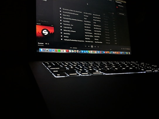A darkly lit scene featuring a laptop with a glowing keyboard and a screen displaying a music streaming application. The interface shows a playlist with visible song titles and album artwork. Application icons are aligned at the bottom of the screen, indicating a computer interface.