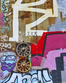 A textured urban wall heavily decorated with graffiti and street art, showcasing a range of abstract shapes, vibrant colors, and two rusty industrial fans embedded into the surface. The graffiti includes stylized letters, abstract forms, and cartoonish characters, including a face with exaggerated features.