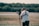 A person operates a professional video camera mounted on a stabilizer, standing in an outdoor setting with blurred greenery in the background. The person is wearing a white t-shirt and a harness, focused on filming.