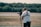 A person operates a professional video camera mounted on a stabilizer, standing in an outdoor setting with blurred greenery in the background. The person is wearing a white t-shirt and a harness, focused on filming.