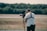 A person operates a professional video camera mounted on a stabilizer, standing in an outdoor setting with blurred greenery in the background. The person is wearing a white t-shirt and a harness, focused on filming.