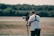 A person operates a professional video camera mounted on a stabilizer, standing in an outdoor setting with blurred greenery in the background. The person is wearing a white t-shirt and a harness, focused on filming.