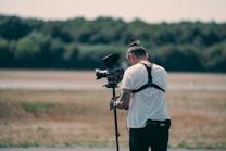 A person operates a professional video camera mounted on a stabilizer, standing in an outdoor setting with blurred greenery in the background. The person is wearing a white t-shirt and a harness, focused on filming.