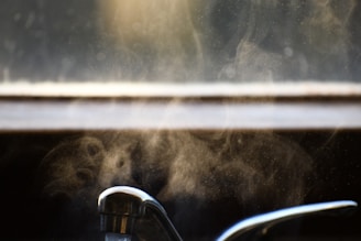 A chrome kitchen faucet is situated below a window with visible steam rising, creating a misty effect. The background appears blurred with light filtering through, enhancing the atmospheric mood.