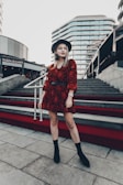 A woman stands confidently on a red-carpeted staircase in an urban setting, wearing a stylish red patterned dress, black boots, and a wide-brimmed hat. Modern architectural buildings serve as the backdrop, while another person is seen in the distance ascending the stairs.