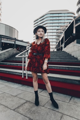 A woman stands confidently on a red-carpeted staircase in an urban setting, wearing a stylish red patterned dress, black boots, and a wide-brimmed hat. Modern architectural buildings serve as the backdrop, while another person is seen in the distance ascending the stairs.