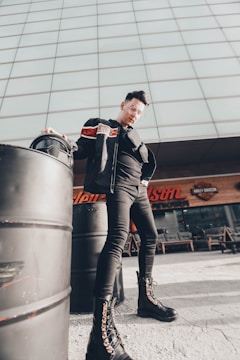 A person is standing confidently next to large metal barrels, wearing a black jacket, sunglasses, and black combat boots with intricate laces. The background features a modern building facade with a sign that includes 'Harley-Davidson'.