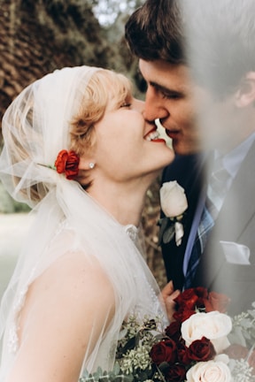 A couple is shown in a close embrace, leaning in for a kiss. The woman, dressed in a white veil with a red flower, holds a bouquet of red and white roses. The background features a blurred natural setting, adding a romantic atmosphere.