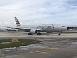 A passenger airplane with the American Airlines logo is positioned on an airport taxiway, featuring overcast skies and various airport markings on the ground.