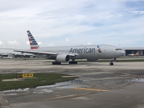 A passenger airplane with the American Airlines logo is positioned on an airport taxiway, featuring overcast skies and various airport markings on the ground.