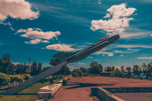 A vintage fighter jet is displayed as a monument, mounted on a large angled support. The setting is an outdoor park with trees lining the background and a blue sky abundant with fluffy white clouds.
